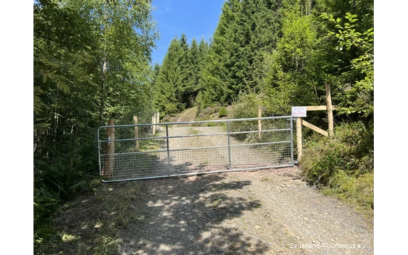 Ein Wanderweg wird von einem metallenen Gatter entzweigeteilt. Mittels eines Schilds wird darum gebeten, das Tor zu schließen.