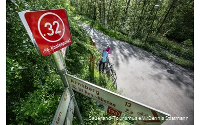 Schild mit einem Knotenpunkt Aufsatz. Im Hintergrund zwei Radfahrer