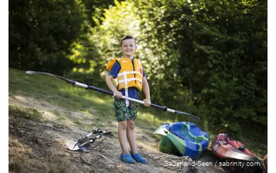 Junge mit Schwimmweste freut sich auf Kanufahrt mit einem Paddel in der Hand.