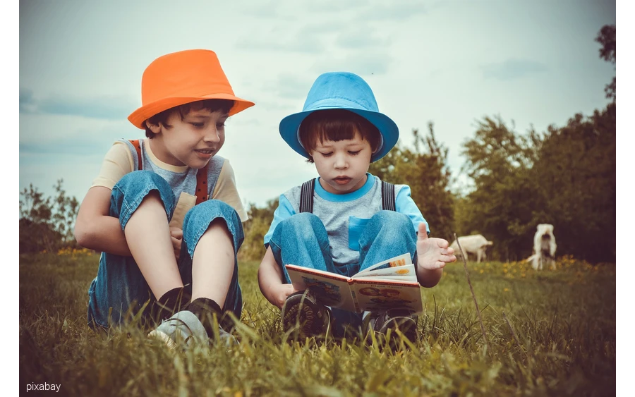 Two boys are sitting in a cow meadow reading a book.
