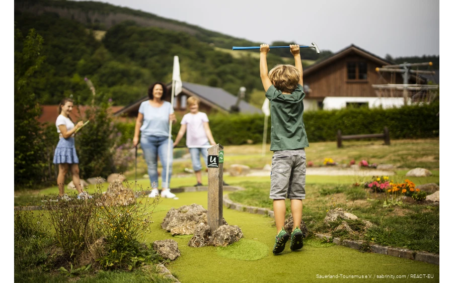 Eine Familie spielt Minigolf. Im Vordergrund freut sich ein Sohn und springt in die Luft.