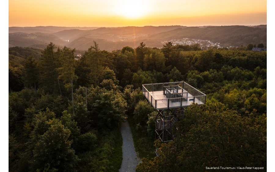 Blick von oben auf den Turm. Im Hintergrund geht die Sonne unter und man sieht verschiedene Ortschaften