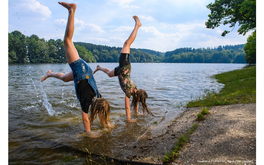 Twee meisjes doen een handstand op de oever van een dam.