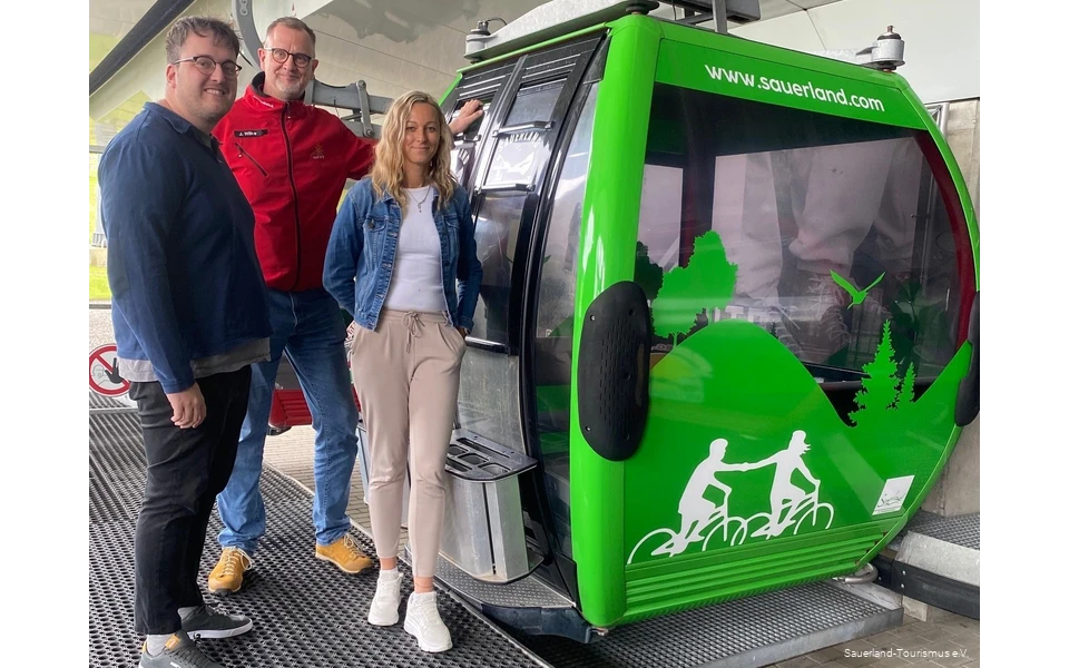 Neben der neu beklebten, grünen Gondel stehen zwei Mitarbeiter vom Sauerland-Tourismus und einer von der Ettelberg-Seilbahn und grinsen.