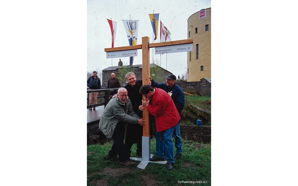 Vier Männer stellen erfreut den ersten Wegweiser des Rothaarsteigs im Rahmen seiner Eröffnung auf. Auf dem Schild sind die Distanzen zu den beiden Start-/Endpunkten des Fernwanderwegs zu sehen.