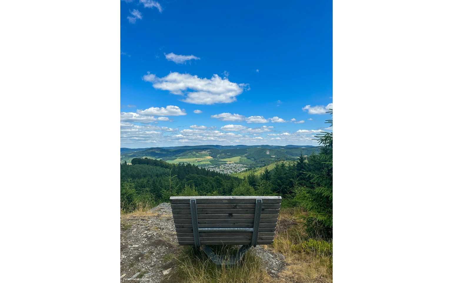 Bank auf einem Felsen mit weiter Aussicht über Wiesen und Wälder und einer Ansammlung von Häusern.