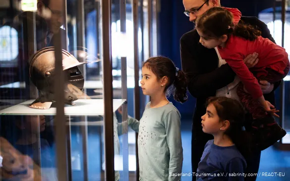 Vater steht mit drei Töchtern vor einer Vitrine in einem Museum. Sie schauen sich einen Ritterhelm an.