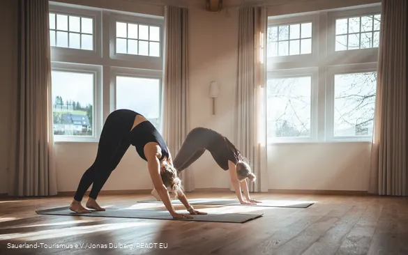 Two women do a yoga exercise in a light-flooded room