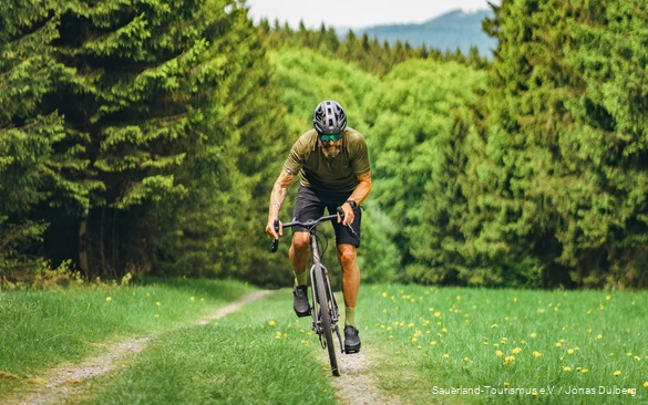 Tätowierter Mann auf einem Gravelbike von vorne. Er fährt einen kleinen Pfad umgeben von Nadelbäumen hoch.