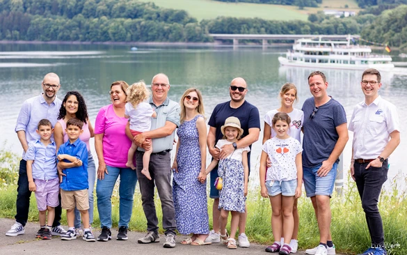Eine lachende Gruppe steht am Ufer des Biggesees mit einem Schiff im Hintergrund