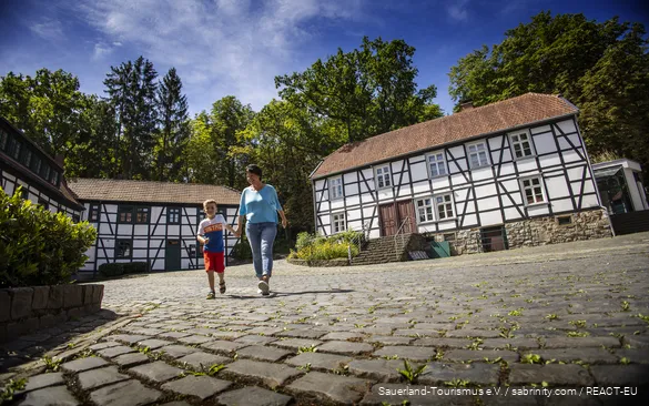 Eine Frau geht mit Kind an der Hand lächelnd über den Hof der Fabrikanlage Maste-Barendorf. Die Gebäude sind alte Fachwerkhäuser.