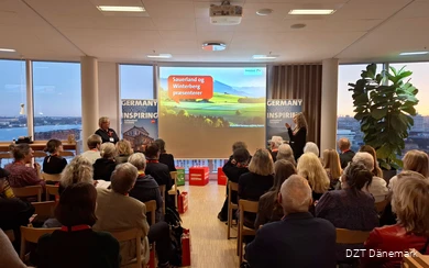 Eine größere Gruppe Menschen schaut auf eine Präsentation. Daneben stehen zwei Frauen, die vortragen.