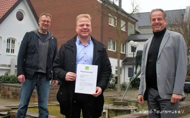 Three men smile, the middle one holds the Sauerland Tourism membership certificate