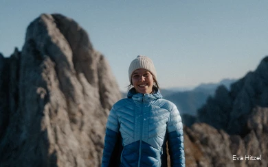 Eva vom Blog Wildwärts grinst mit blauer Daunenjacke und Mütze bekleidet in die Kamera. Im Hinzergrund sind schroffe Berge zu sehen.