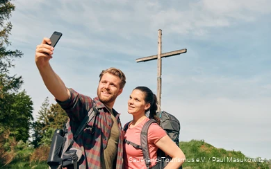 Ein Pärchen in Wanderausrüstung posiert für ein Selfie vor einem Gipfelkreuz.