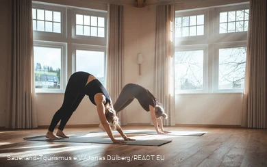 Two women do a yoga exercise in a light-flooded room