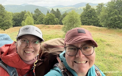 Anne Abendroth en haar vriendin grijnzen in de camera tijdens een van hun wandeltochten. Ze dragen outdoorkleding, een Kappe en een wandelrugzak.