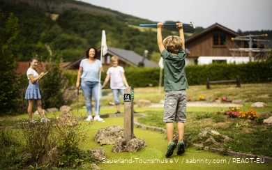 Een gezin speelt minigolf. Op de voorgrond is een zoon blij en springt in de lucht.