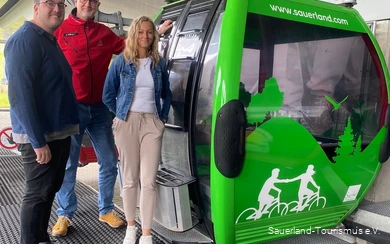 Neben der neu beklebten, grünen Gondel stehen zwei Mitarbeiter vom Sauerland-Tourismus und einer von der Ettelberg-Seilbahn und grinsen.