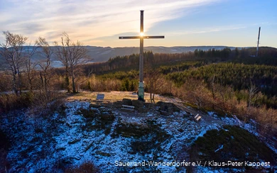 Landschaftsaufnahme von Gipfelkreuz auf dem Olsberg beim Sonnenaufgang. Es liegt eine dünne Schicht Schnee.