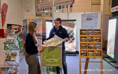 Ein Mitarbeiter des Sauerland-Tourismus berät eine Besucherin der Fahrradmesse in Essen an seinem Stand. Er hält ihr eine Karte des Sauerlands hin.