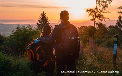 Mann und Frau stehen Arm in Arm mit dem Rücken zur Kamera und schauen sich einen farbenfrohen Sonnenuntergang von einem Aussichtspunkt aus an. Sie tragen Rucksäcke und Outdoorkleidung.