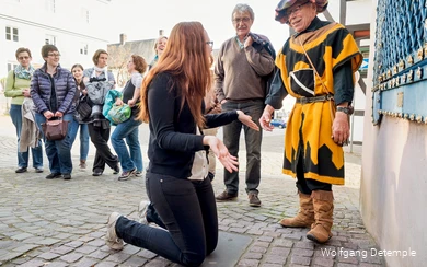 Im Vordergrund kniet eine Frau vor einer Mauer, den Blick auf einen verkleideten Mann gerichtet. Im Hintergrund steht der Rest der Gruppe und amüsiert sich.