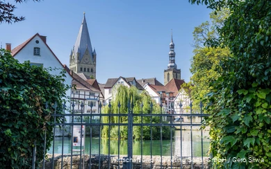 in the foreground is a pond in the center of Soest, in the background you can see half-timbered houses and two church towers under a bright blue sky in summer