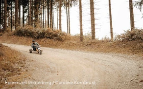Eine Person fährt mit einem Mountaincart, Fahrzeug mit drei Rädern und einem Lenker, durch eine Kurve einen Waldweg hinab.