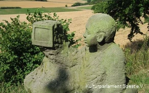 Human stone sculpture looks into a television surrounded by distant views.