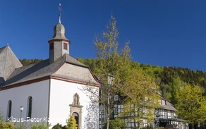 Eine Kirche vor blauem Himmel neben einem Fachwerkhaus, umgeben von Bäumen.