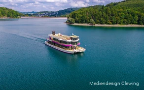 Das Personenschiff EMS Hennesee mit pinken Farbakzenten und einem großen Sonnendeck fährt über den Hennesee. Im Hintergrund ist die Staumauer und ein Teil Meschedes zu sehen.