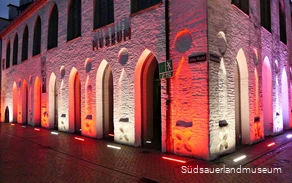 Beleuchtete Fassade des Südsauerlandmuseums in rot und weiß. An der Mauer steht die Aufschrift Museum.