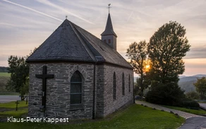 Eine steinerne Kapelle umgeben von einer grünen Wioese mit der untergehenden Sonne im Hintergrund.