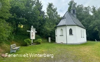 Kapelle mit einer Jesusfigur am Kreuz und einer Bank, umgeben von Bäumen.