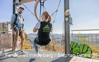 Eine Frau sitzt in einem Sitzgurt, welcher an einer Metallschiene hängt auf einer Startplattform.