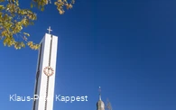 Eine Kirche neben einem hohen Kirchturm mit Turmuhr vor blauem Himmel.
