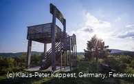 Ein Aussichtsturm in Form eines großen Holzstuhles mit einer Treppe, die hinauf zur Aussichtsplattform führt, inmitten von Bäumen und Wiesen.