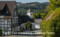 Einige Häuser und eine Kirche an einer Straße mit Bergen und Wäldern im Hintergrund.