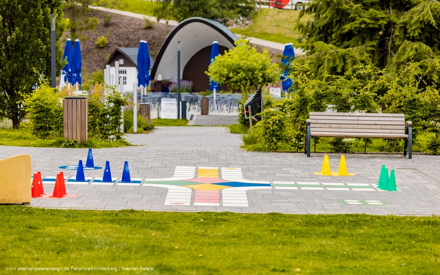 Im Vordergrund ein großes, ins Pflaster integriertes, Mensch ärgere dich nicht Spiel mit der Musikmuschel im Hintergrund.