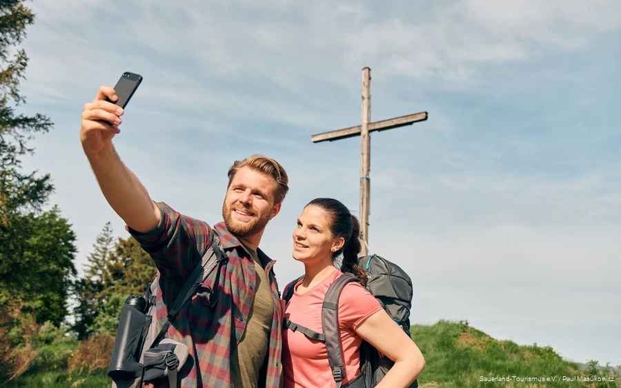 Ein Pärchen in Wanderausrüstung posiert für ein Selfie vor einem Gipfelkreuz.