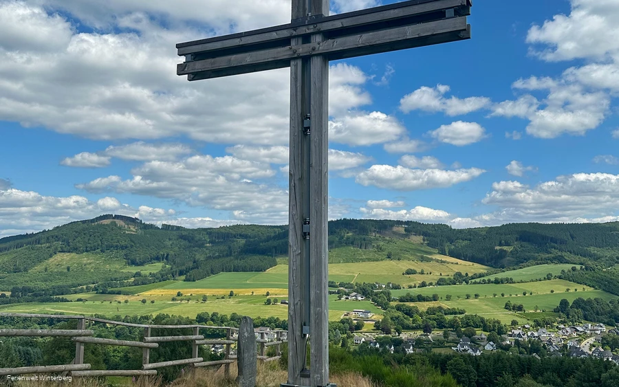 Ein großes Holzkreuz, dahinter eine weite Aussicht über Wiesen, Wälder und Berge.