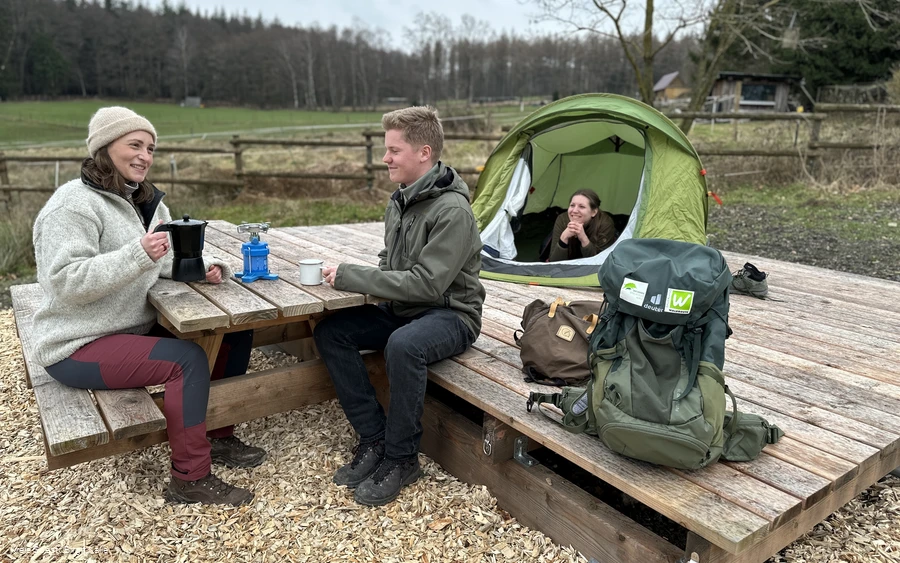 Twee wandelaars zitten aan een tafel op een trekpleister in het Arsnbergbos. Op de achtergrond staat een tent met een vrouw die naar buiten kijkt.