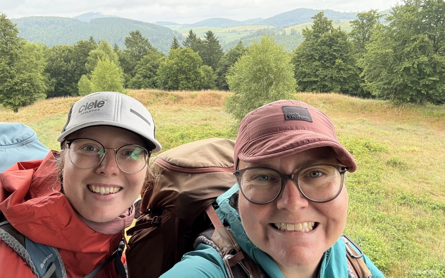 Anne Abendroth en haar vriendin grijnzen in de camera tijdens een van hun wandeltochten. Ze dragen outdoorkleding, een Kappe en een wandelrugzak.