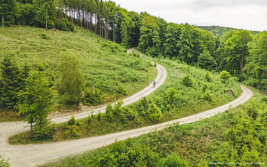 Luftaufnahme von einem Mann auf einem Gravelbike durch grüne Landschaft. Der Weg macht eine 180-Grad-Kurve.
