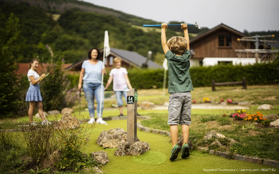 A family playing mini golf. In the foreground, a son is happy and jumps into the air.