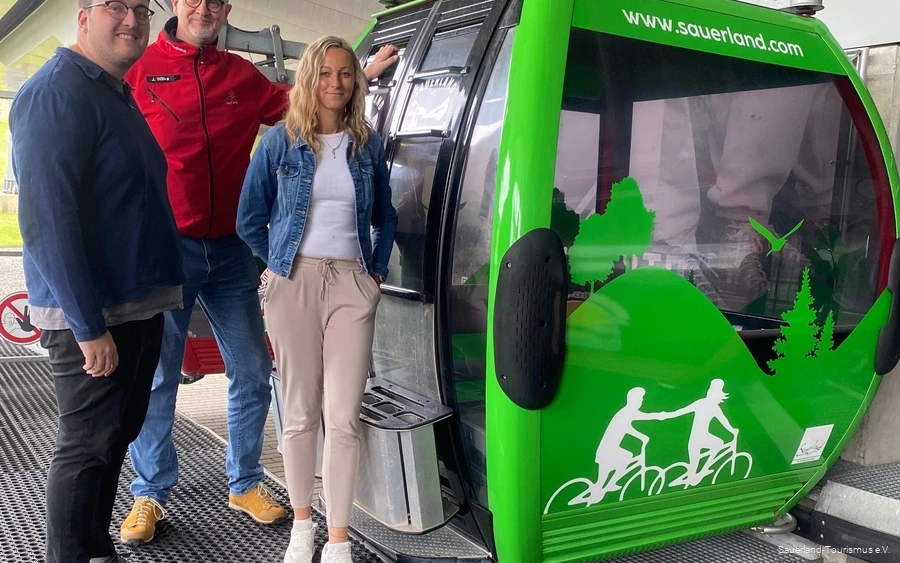 Neben der neu beklebten, grünen Gondel stehen zwei Mitarbeiter vom Sauerland-Tourismus und einer von der Ettelberg-Seilbahn und grinsen.