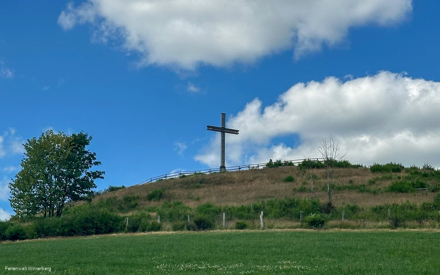 Ein Kreuz auf einem begrünten Hügel vor blaum Himmel mit Wolken.