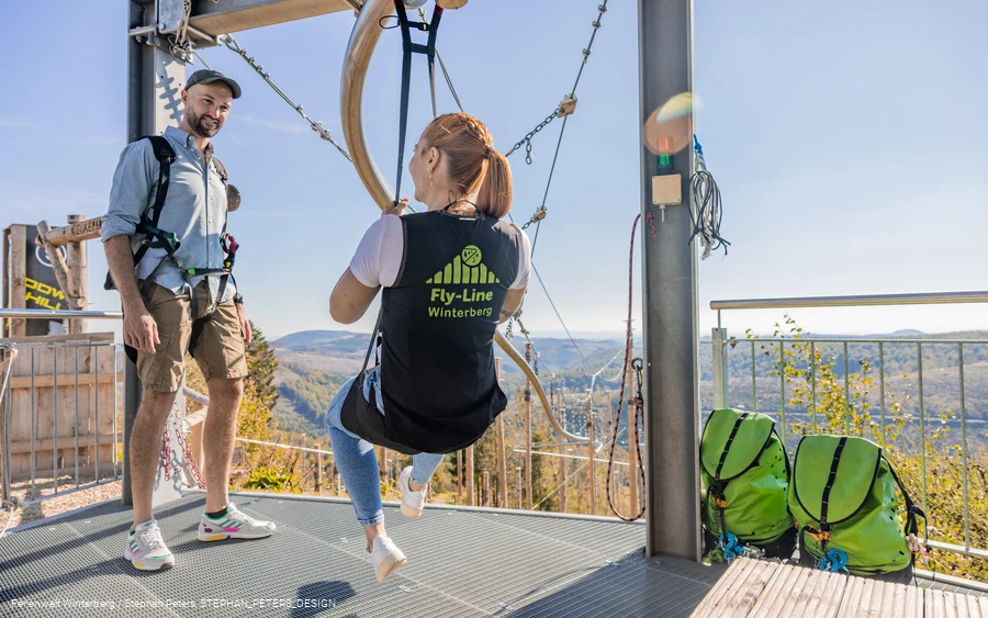 Eine Frau sitzt in einem Sitzgurt, welcher an einer Metallschiene hängt auf einer Startplattform.