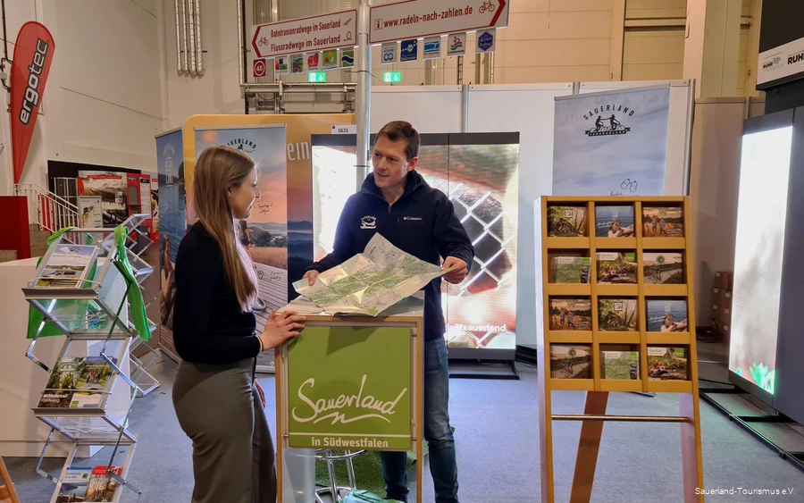 Ein Mitarbeiter des Sauerland-Tourismus berät eine Besucherin der Fahrradmesse in Essen an seinem Stand. Er hält ihr eine Karte des Sauerlands hin.